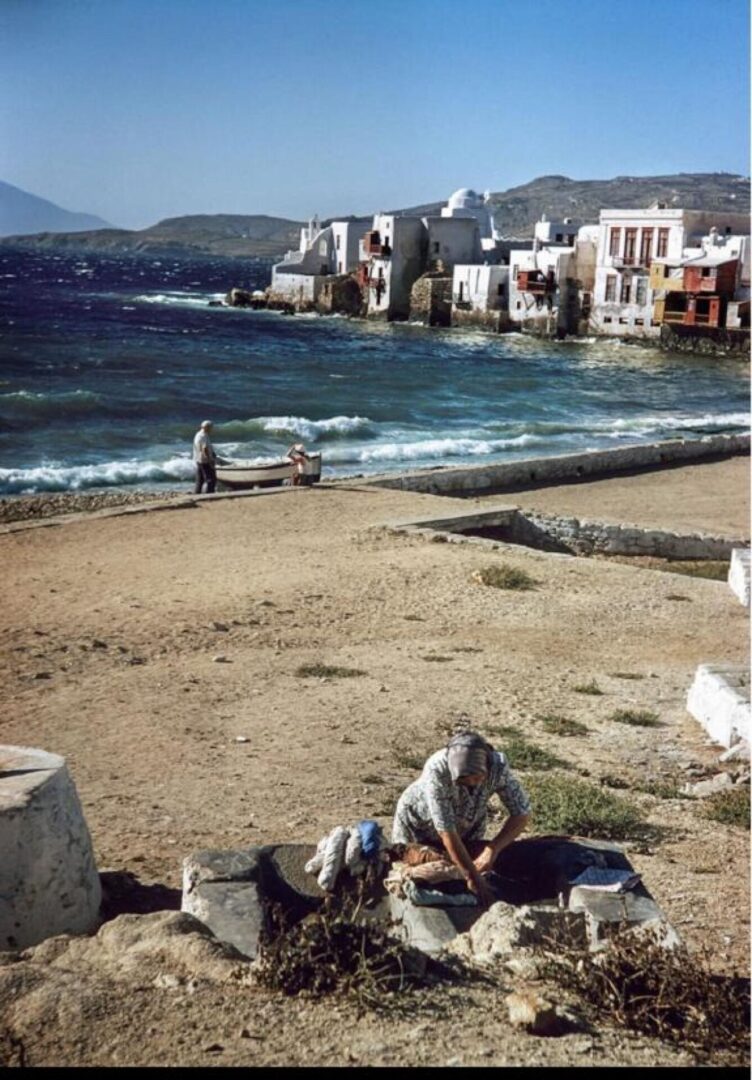 Μύκονος, 1957. Ανεμοδαρμένη ημέρα του Ιουλίου. Μια γυναίκα πλένει τα ρούχα της με θέα τη Μικρή Βενετία ενώ στην παραλία ένας άντρας φροντίζει τη βάρκα του